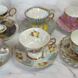Könnte beinhalten: Eine Sammlung von Vintage-Teetassen und Untertassen in verschiedenen Designs. Die Teetassen weisen goldene Verzierungen, florale Muster und einzigartige Formen auf. Untertassen haben gewellte Ränder und ergänzende Designs. Die Farbpalette umfasst Rosa, Weiß, Gold und Schwarz.
