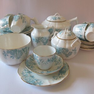 May include: A complete vintage tea set in white with a teal floral pattern and gold trim. The set includes a teapot, sugar bowl, creamer, cups, saucers, and a large bowl. The edges of the pieces are trimmed with gold.