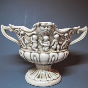 Peut inclure: Pot à fleurs en céramique crème avec une base à piédestal et deux poignées. Le pot présente un motif en relief détaillé de chérubins, un visage barbu et des éléments décoratifs. Le style général rappelle l'art classique.