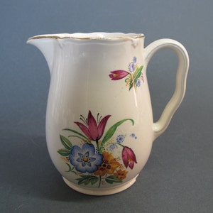 Puede incluir: Una jarra de cerámica blanca con un diseño floral. La jarra tiene un borde festoneado y un asa. Las flores son rosas, azules, amarillas y verdes.