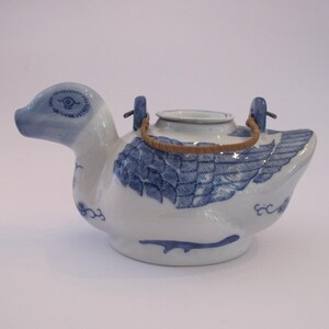 May include: A blue and white porcelain teapot shaped like a duck with a brown woven handle. The teapot has a blue and white design of a duck with feathers and a stylized eye.