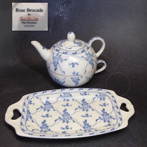 Puede incluir: Una tetera y una bandeja a juego de porcelana azul y blanca con un diseño floral. La tetera es de doble pared con una taza en la parte superior. La bandeja tiene dos asas. La etiqueta dice "Rose Brocade by Skye McChie Fine Porcelain Made in China".