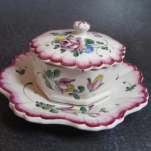 May include: A white ceramic butter dish with a lid, featuring a floral design in pink, yellow, blue, and green. The dish and lid have a scalloped edge with a pink trim, and the dish sits on a matching plate.