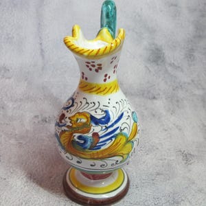 May include: A small, white ceramic pitcher with a green handle. The pitcher is decorated with a colorful, stylized bird design in shades of blue, yellow, red, and green. The base of the pitcher is decorated with a yellow and red band.