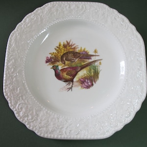 Peut inclure: Assiette carrée en céramique blanche avec une bordure en relief. Le centre de l'assiette présente une illustration colorée de deux faisans dans un champ herbeux.