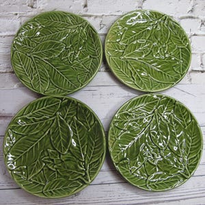 Peut inclure: Quatre assiettes rondes en céramique verte avec un motif de feuilles en relief. Les assiettes ont une finition brillante. Le motif de feuilles recouvre toute la surface de chaque assiette, créant une apparence texturée. Les assiettes sont disposées en carré.
