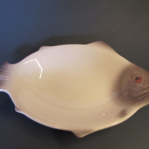 May include: White ceramic fish-shaped serving dish with a grey and white painted fish design. The dish has a red painted eye.