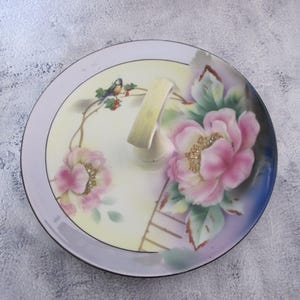 May include: A vintage porcelain serving plate with a handle. The plate features a floral design with pink roses, green leaves, and a small blue bird. The plate has a white background with a light purple border.