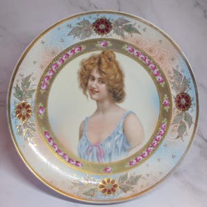 May include: A round porcelain decorative plate featuring a central portrait of a person with wavy hair wearing a light blue top. The border is decorated with a gold band, pink rose garlands, and four floral motifs in red and green.