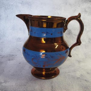 May include: A copper pitcher with blue and gold painted designs. The pitcher has a wide base and a rounded body. The handle is attached to the side of the pitcher.
