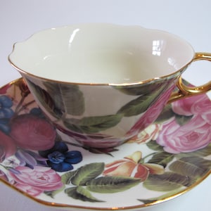 May include: A white teacup and saucer with a gold rim. The cup and saucer are decorated with a floral pattern of pink, red, and green roses.