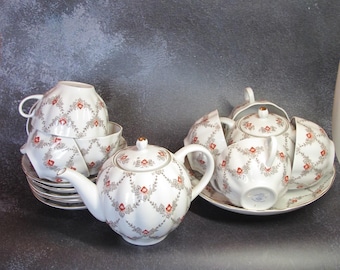 May include: A white porcelain tea set with a floral pattern in red and gray. The set includes a teapot, cups, saucers, and a sugar bowl. The teapot has a gold-colored knob on the lid. The cups and saucers are stacked.