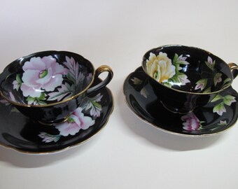 Puede incluir: Dos tazas de té negras y platillos con ribete dorado. Las tazas presentan diseños florales en rosa, amarillo y verde.