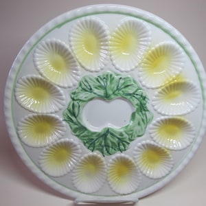 Puede incluir: Un plato de servir de cerámica blanca con 12 secciones acanaladas amarillas y blancas para contener huevos u otros bocadillos pequeños. El centro del plato presenta un diseño de hojas verdes.