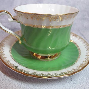 May include: A vintage teacup and saucer set. The teacup is green with a white rim and gold trim. The saucer is white with a green center and gold trim. The handle of the teacup is white with gold accents. A classic tea set.