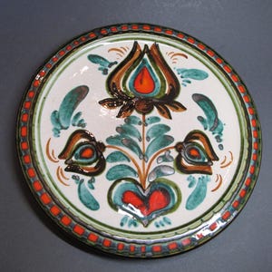 May include: Round ceramic plate with a white base, featuring a central floral design in brown, orange, and green. The border has a red and green geometric pattern. Decorative tableware.