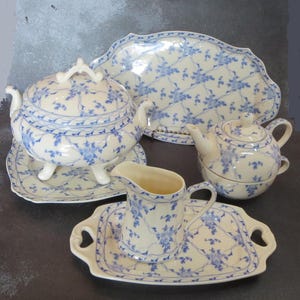 May include: A cream porcelain tea set with a blue floral design. The set includes a teapot, a lidded serving dish, a creamer, and serving trays. The items are arranged on a dark surface.