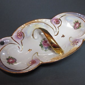 May include: Oval porcelain serving dish with a gold handle. The dish has a white base with pink and purple floral designs, gold trim, and a pearlescent sheen. Decorative holes are on the rim.