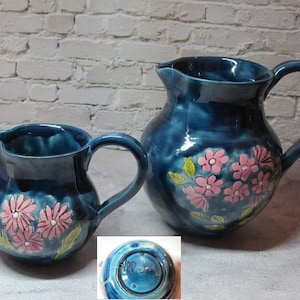 May include: Two blue ceramic jugs with pink floral designs. The smaller jug has a blue and white base with the word "Rose" written on it.