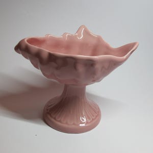 May include: A pink ceramic seashell-shaped bowl with a pedestal base. The bowl is designed to resemble a seashell with a ruffled edge.