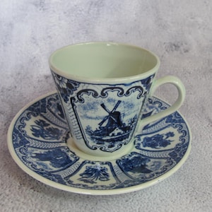 Puede incluir: Un juego de taza y platillo blanco con un diseño de molino de viento azul. La taza y el platillo tienen un patrón azul detallado en los bordes. La taza tiene un asa curva.