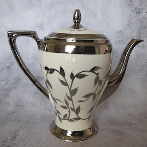 Peut inclure: Une théière en céramique blanche avec un motif de feuilles argentées et une bordure argentée. La théière a un couvercle avec un bouton argenté.