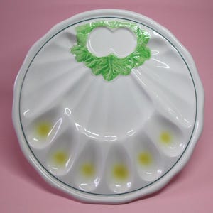 Könnte beinhalten: Weiße Keramik-Eierplatte mit einem grünen Blattdesign in der Mitte. Die Platte hat acht einzelne Abschnitte zum Halten von Eiern, jeder mit einer gelben Mitte.