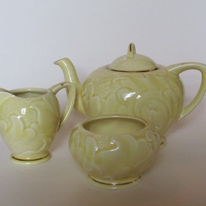 Peut inclure: Service à thé en porcelaine jaune pâle, comprenant une théière avec couvercle, un pot à crème et un sucrier. L'ensemble présente un motif floral en relief et des bordures dorées. La théière mesure environ 15 cm de haut.