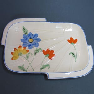 以下が含まれることがあります： 青い縁取りと花柄の白いセラミック製のサービングディッシュ。花は青、オレンジ、黄色の色合いで描かれています。