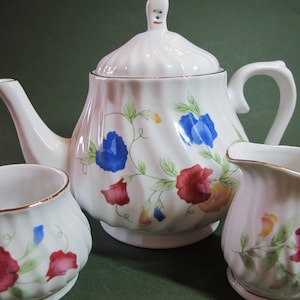 Peut inclure: Une théière, un pot à crème et un sucrier blancs avec un motif floral composé de fleurs rouges, bleues et jaunes. La théière a un couvercle et une bordure dorée. Le pot à crème et le sucrier ont également une bordure dorée.
