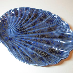 Puede incluir: Un plato de cerámica azul en forma de concha con un patrón moteado. El plato tiene un borde festoneado y una superficie texturizada con líneas radiales. El color es azul oscuro con detalles en azul oscuro y negro.