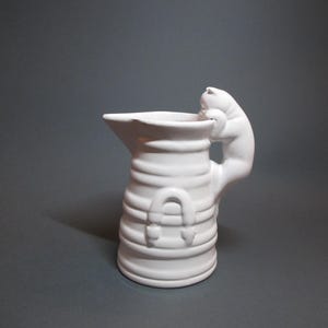 Peut inclure: Pichet en céramique blanche avec une anse en forme de chat. Le pichet a une surface texturée et un motif en relief de fer à cheval sur le devant.