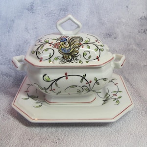 May include: A white ceramic soup tureen with a matching lid and saucer. The tureen features a rooster design and floral accents in green, red, and black. The lid has a handle, and the tureen has side handles.