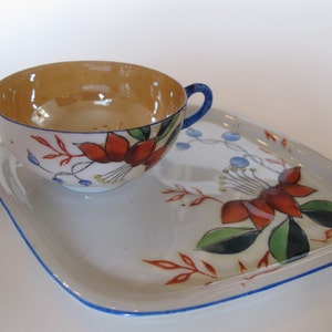 Könnte beinhalten: Ein weißes Porzellan-Teetassen- und Untertassen-Set mit einem Blumendesign. Die Tasse hat einen goldenen Rand und die Untertasse einen blauen Rand. Das Blumendesign zeigt rote, orange, gelbe, grüne und blaue Blumen.