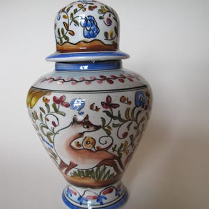 Könnte beinhalten: Dekorative Keramikvase mit Deckel, handbemalt. Sie zeigt ein Reh, Blumen und Blätter in Braun-, Blau-, Rot- und Gelbtönen auf weißem Hintergrund. Die Vase hat einen blauen Sockel und Rand.