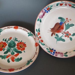 May include: Two white ceramic plates with hand-painted floral designs. One plate features a red bird with green leaves and red flowers. The other plate has red and green flowers.