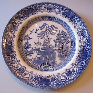 May include: A blue and white porcelain dinner plate with a willow pattern design. The plate features a traditional Chinese landscape scene with a bridge, trees, and a pagoda.