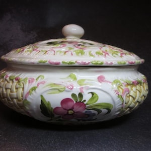 May include: A white ceramic lidded bowl with a floral design. The bowl has a woven basket pattern around the base and is decorated with pink and green floral accents. The lid has a small, round knob.