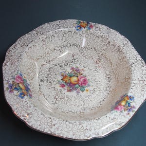 May include: A white ceramic bowl with a gold and floral design. The bowl has a scalloped edge and features a central floral motif with pink, yellow, and blue flowers.