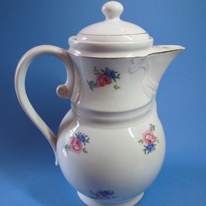 Peut inclure: Cafetière en céramique blanche avec un motif floral composé de fleurs roses et bleues. La cafetière a un couvercle avec une bordure dorée et une poignée incurvée.
