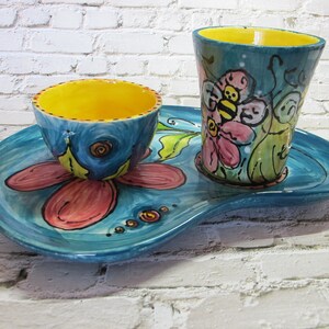 Puede incluir: Una bandeja de cerámica azul con un diseño de flor rosa y detalles amarillos. Una taza de cerámica azul con un borde amarillo y un diseño de abeja y flor se encuentra en la bandeja. Un tazón de cerámica azul más pequeño con un borde amarillo se encuentra en la bandeja.