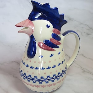May include: A ceramic pitcher in the shape of a rooster, featuring a white body with blue and pink accents. The rooster has a blue comb, pink beak, and blue wattle. The pitcher is decorated with pink dots and blue floral patterns.