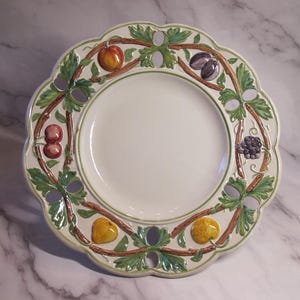 Puede incluir: Plato de cerámica blanca con borde festoneado, decorado con un diseño de vid verde y marrón con varias frutas. Las frutas incluyen melocotones, ciruelas, cerezas y moras. El plato tiene un borde verde.