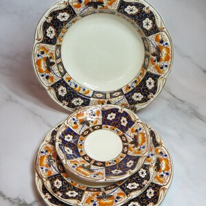May include: A collection of vintage ceramic plates. The plates have a cream-coloured centre and a decorative border with orange, navy blue, and white floral patterns. Scalloped edges and gold trim complete the design.