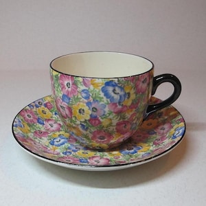 Op de afbeelding: Een bloemetjes theekopje en schotelset met een zwarte rand. Het kopje en de schotel zijn bedekt met een patroon van roze, gele, blauwe en groene bloemen.
