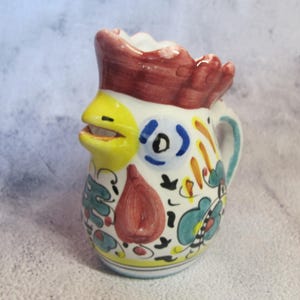 May include: A ceramic pitcher with a rooster design. The pitcher features a yellow beak, a red comb, and colorful floral patterns. The handle is light blue. The pitcher is approximately 5 inches tall.