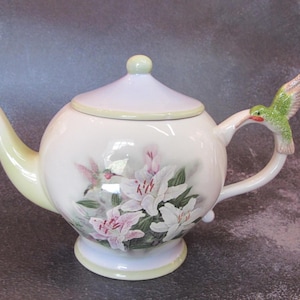 Puede incluir: Una tetera de cerámica blanca con tapa y asa de colibrí. La tetera está decorada con un diseño floral con lirios blancos y rosas. El pico y la base tienen un borde amarillo pálido.