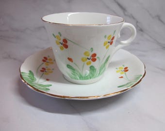 May include: A white teacup and saucer with a floral pattern. The cup and saucer have a gold trim around the edge. The flowers are yellow, orange, and green.