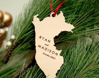 May include: A light-coloured wooden ornament shaped like the state of New Jersey, featuring the engraved text RYAN and MADISON with October 2023 below it. The ornament is suspended by a thin red ribbon through a hole at the top of the wooden cutout.
