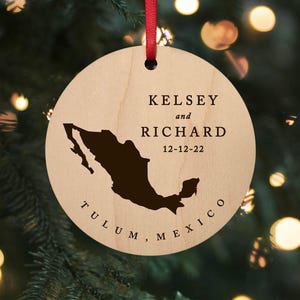 May include: A round, light-colored wooden ornament hanging from a red ribbon. It features a dark brown silhouette of Mexico, the names KELSEY and RICHARD, the date 12-12-22, and the text TULUM, MEXICO printed in a dark, serif font on the front.